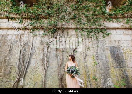 Schöne Braut in einem Pastellkleid mit einem luxuriösen Blumenstrauß steht vor dem Hintergrund einer Steinwand, die mit g umschlungen ist Stockfoto