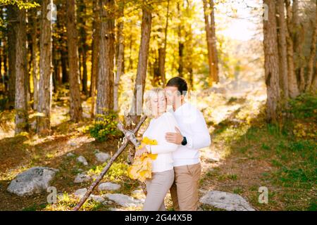 Mann küsst und umarmt lächelnde Frau an den Schultern. Die Frau hat gelbe Blätter in ihren Händen. Weitwinkel Stockfoto