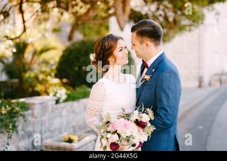 Frisch verheiratetes Paar lächelt sich zärtlich vor dem Hintergrund des Grüns an. Die Braut hält einen schönen Strauß in ihren Händen Stockfoto