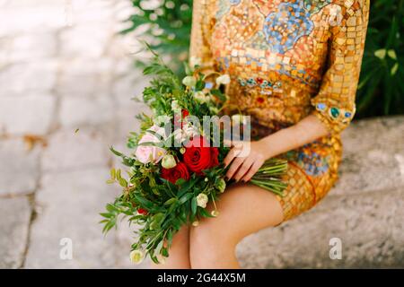 Eine Braut in einem ungewöhnlichen goldenen Kleid mit Ikone sitzt und hält einen Brautstrauß aus weißen und roten Rosen in der Hand, Nahaufnahme Stockfoto