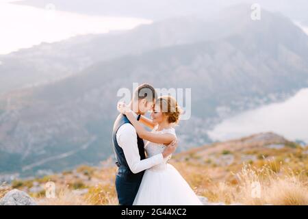 Die Braut und der Bräutigam stehen auf dem Gipfel des Mount Lovcen mit Blick auf die Bucht von Kotor und umarmen sich liebevoll aus nächster Nähe Stockfoto