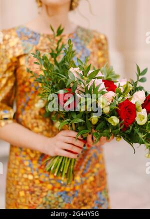 Eine Braut in einem ungewöhnlichen goldenen Kleid hält einen Brautstrauß aus weißen und roten Rosen in den Händen, aus nächster Nähe Stockfoto