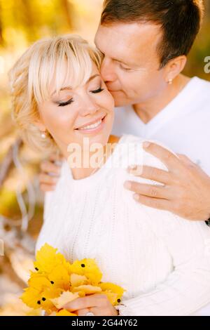 Mann küsst und umarmt lächelnde Frau an den Schultern. Die Frau hat gelbe Blätter in ihren Händen Stockfoto