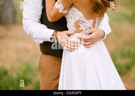 Der Bräutigam hält seine Hände in einem schönen, bestickten weißen Kleid an der Taille der Braut. Die Braut steht mit dem Rücken Stockfoto