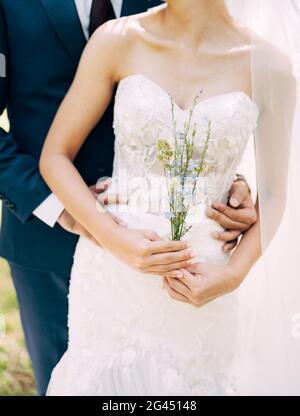 Bräutigam in einem blauen Anzug Umarmungen von hinten Braut in einem weißen off-the-shoulders Kleid. Braut hält Wildblumen in ihren Händen Stockfoto