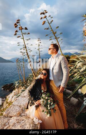 Die Braut in einer Lederjacke und Sonnenbrille mit einem Blumenstrauß sitzt auf einem Stein und lehnt sich an den neben h stehenden Bräutigam Stockfoto