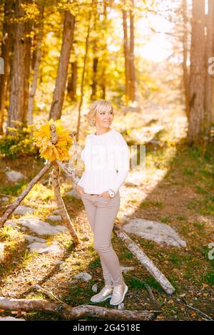 Lächelnde Frau in einem weißen Pullover steht neben einem Baum. An den Zweigen hängt ein Kranz aus gelben Blättern Stockfoto