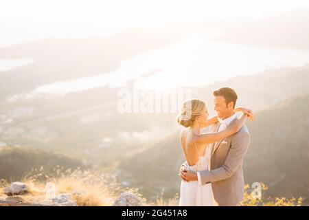 Die Braut und der Bräutigam stehen auf dem Gipfel des Mount Lovcen mit Blick auf die Bucht von Kotor, lächeln und umarmen sie zärtlich Stockfoto