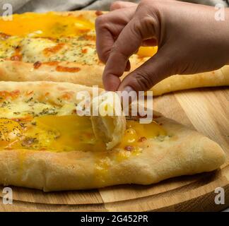 Gebackene adjarian khachapuri mit dem ganzen Eigelb auf einem Holzbrett, schlägt die Hand ein Stück Brot in ein ausgetretenes Eigelb Stockfoto