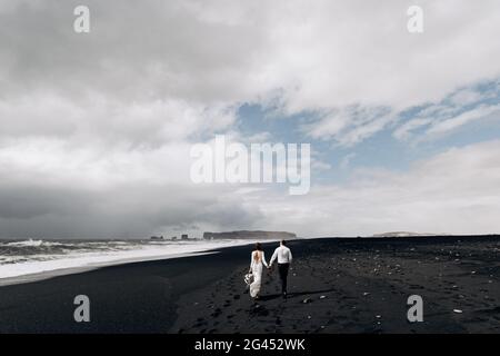 Ein Hochzeitspaar läuft am schwarzen Strand von Vic entlang. Sandstrand mit schwarzem Sand am Ufer des Atlantischen Ozeans. Braut Stockfoto