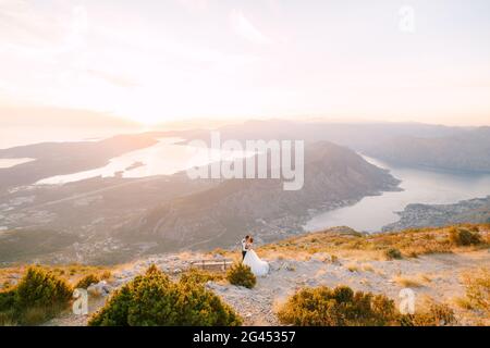 Die Braut und der Bräutigam stehen auf dem Gipfel des Mount Lovcen mit Blick auf die Bucht von Kotor in der Nähe einer Holzbank, küssen und umarmen sie zärtlich Stockfoto