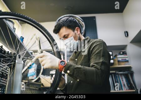 Mechaniker repariert Fahrrad in der Werkstatt trägt covid 19 Maske. Arbeiter des Fahrradspeichers repariert Fahrrad während der Coronavirus-Quarantäne in Stockfoto