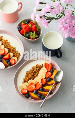 Leckeres gesundes Frühstück, Müsli mit Joghurt und süßen Früchten, Banane und Erdbeeren, Blaubeeren und Pfirsiche, Milch im Becher und Blumen Stockfoto