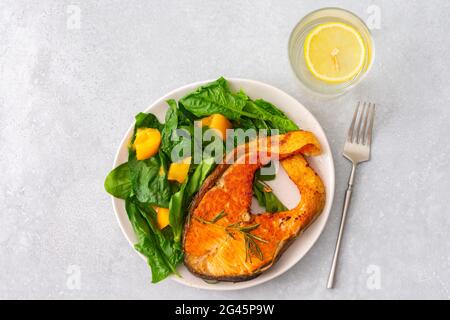 Gebratenes Lachssteak mit Spinat und gelben Tomaten auf einem Teller auf grauem Grund, ein Glas Wasser mit Zitrone, ein herzhaftes Mittagessen mit gesundem rotem Fisch Stockfoto