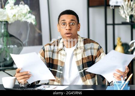 Schockiert staunte Latino Kerl, in einer modernen Kleidung und Brille, Manager oder Büroangestellte, sitzen am Tisch, Dokumente, Charts in den Händen, Blick direkt in die Kamera überrascht Stockfoto