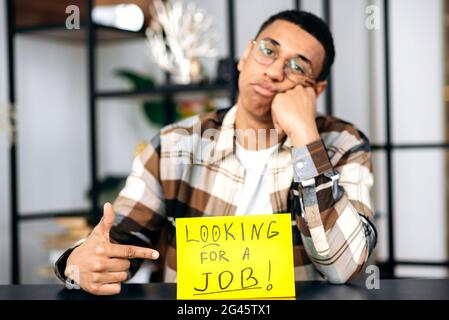 Unverschämter hispanischer trauriger arbeitsloser Typ, Student oder Freiberufler, mit Brille und in legerem Hemd, sitzt an seinem Schreibtisch, zeigt auf ein Schild, DAS NACH ARBEIT sucht, schaut aufgeregt auf die Kamera und hofft, einen Job zu bekommen Stockfoto