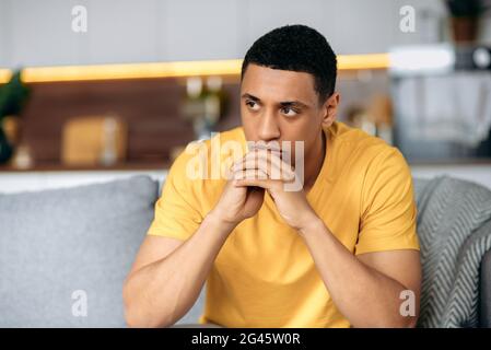 Enttäuscht unglücklicher hispanischer Tausendjähriger in einem gelben T-Shirt, der auf dem Sofa im Wohnzimmer sitzt, hat persönliche Probleme, Versagen bei der Arbeit, Depressionen, schlechte Laune, trauriger junger Mann ist einsam Stockfoto