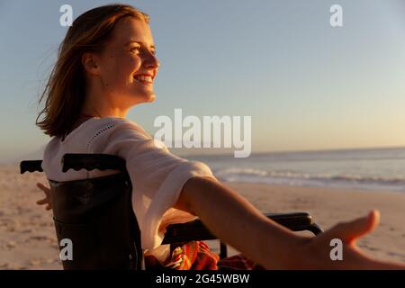 Lächelnde Frau im Rollstuhl mit weit geöffneten Armen am Strand Stockfoto