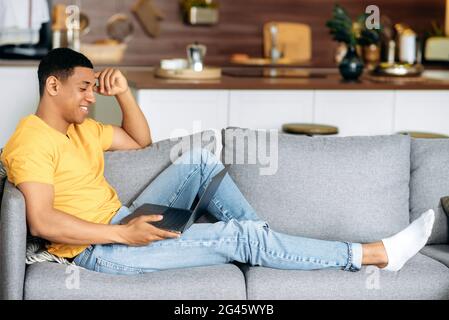 Positiv attraktiver hispanischer Mann, der zu Hause im Wohnzimmer auf einem Sofa sitzt, einen Laptop benutzt, im Internet surft, online in sozialen Netzwerken chattet, online einkauft, Laptop anschaut und lächelt Stockfoto