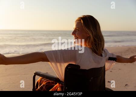 Lächelnde Frau im Rollstuhl mit weit geöffneten Armen am Strand Stockfoto