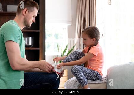 Kaukasisches Mädchen sitzt auf einer Couch, während ihr Vater hilft ihr, Schuhe anzuziehen Stockfoto