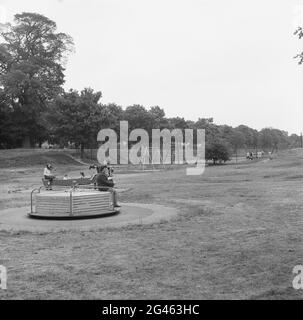 1969, historische, Kinder auf einem Spielplatz in einem öffentlichen Park, zeigt die Spielgeräte des Tages; ein Karussell oder ein Karussell, ein Metall-Spinnrad, angetrieben von den spielenden Kindern und ein Metall-Schaukelpferd, alle später verboten, als zu gefährlich angesehen, England, Großbritannien. Stockfoto