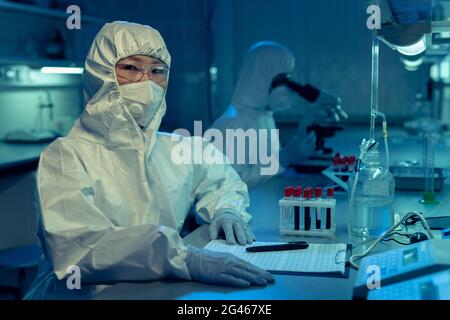 Junge Forscherin in Overalls und anderen schützenden Arbeitskleidung im Labor sitzen Stockfoto