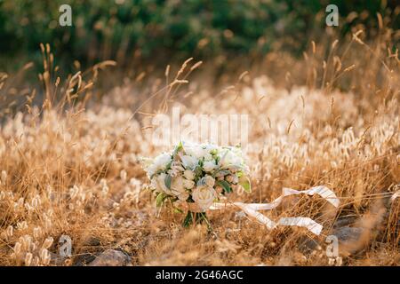 Brautstrauß aus weißen Rosen, Calla-Lilien, Geißelblumen, stachys und weißen Bändern auf dem Boden im Gras Stockfoto