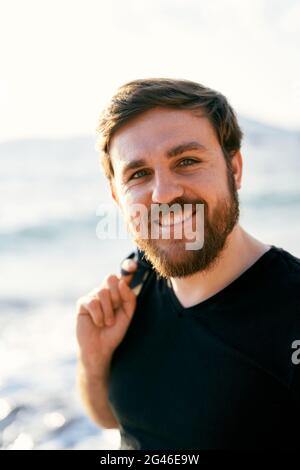 Lächelnder Mann in einer Jeansjacke über der Schulter steht vor dem Hintergrund des Meeres. Nahaufnahme. Hochformat Stockfoto