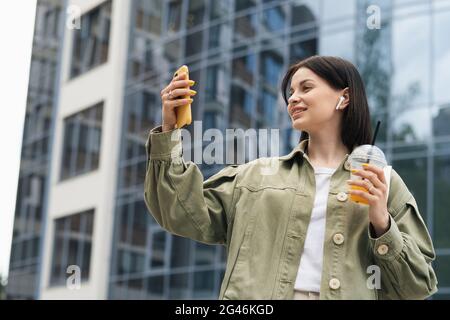 Glückliche junge Frau, die sich mit einem Cocktailgetränk in der Hand mit ihrem Mobiltelefon fotografiert, kaukasische Frau, die Selfie mit dem Smartphone auf der Straße macht Stockfoto