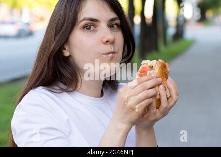 Brünette Frau überfrisst auf einem Burger auf der Straße. Völlerei, überschüssige Kalorien und Bulimie. Stockfoto