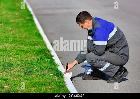 Ein Arbeiter in Overalls malt an einem Sommertag die Bordsteinkante mit weißer Farbe. Städtischer Dienst, Verbesserung der Territorien. Stockfoto