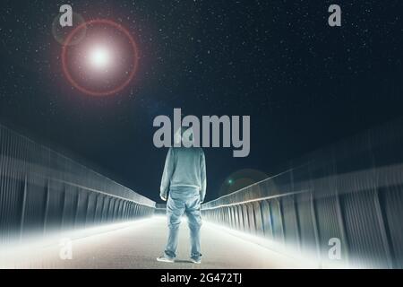Eine geheimnisvolle Kapuzenfigur, die auf einem Pfad steht. Silhouetten gegen helle Lichter in der Nacht. Blick auf ein helles UFO am Himmel Stockfoto