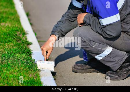 Ein Arbeiter in Overalls malt an einem Sommertag eine Grenze mit weißer Farbe. Städtische Dienstleistungen, Landschaftsbau. Stockfoto