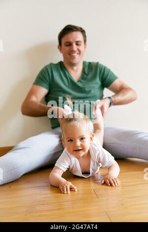 Lächelnder Vater sitzt auf dem Boden und zieht das kriechende niedliche Baby an den Beinen Stockfoto