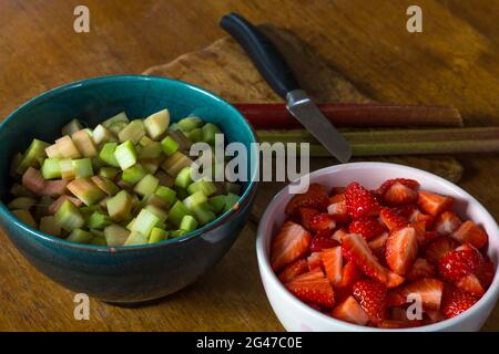 Frische Erdbeeren und Rhabarber in Schüsseln auf der Oberfläche eines Tisches zusammen mit einem Messer, einem Schneidebrett und zwei Rhabarberstielen in Stücke geschnitten Stockfoto