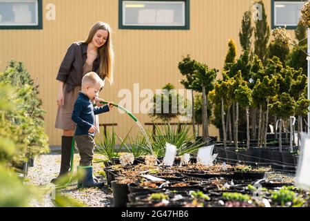 Der kleine Sohn hilft der Mutter, die Töpfe der Pflanzen im Gartencenter zu bewässern Stockfoto