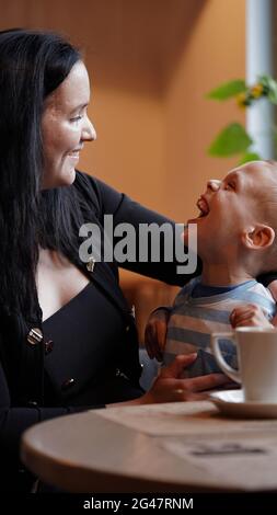 Nahaufnahme Porträt eines kleinen Jungen mit besonderen Bedürfnissen und Mutter lachen an einem Tisch in einem Café, Lifestyle. Die Liebe der Mutter zu ihrem Kind, Inklusion.Happy disab Stockfoto