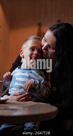 Nahaufnahme Porträt eines kleinen Jungen mit besonderen Bedürfnissen und Mutter lachen an einem Tisch in einem Café, Lifestyle. Die Liebe der Mutter zu ihrem Kind, Inklusion.Happy disab Stockfoto