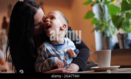 Nahaufnahme Porträt eines kleinen Jungen mit besonderen Bedürfnissen und Mutter lachen an einem Tisch in einem Café, Lifestyle. Die Liebe der Mutter zu ihrem Kind, Inklusion.Happy disab Stockfoto