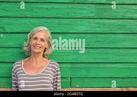 Lächelnde ältere Frau, die gegen die grüne Wand steht Stockfoto