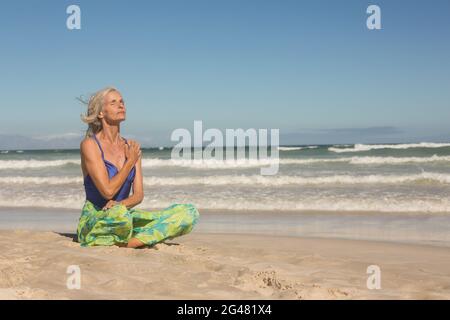 Ältere Frau meditiert, während sie am Ufer gegen den klaren Himmel sitzt Stockfoto