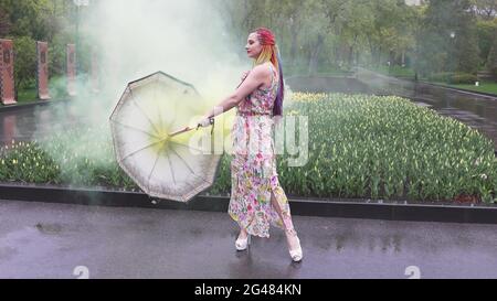 Entzückendes Mädchen in einem Kleid mit einem Blumendruck mit Regenbogen Zöpfen und Make-up. Tanzt mit einem Regenschirm, der sich in gelbem Rauch vor der Kulisse von FL Stockfoto