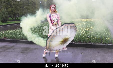 Entzückendes Mädchen in einem Kleid mit einem Blumendruck mit Regenbogen Zöpfen und Make-up. Tanzt mit einem Regenschirm, der sich in gelbem Rauch vor der Kulisse von FL Stockfoto