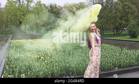 Entzückendes Mädchen in einem Kleid mit einem Blumendruck mit Regenbogen Zöpfen und Make-up. Tanzt mit einem Regenschirm, der sich in gelbem Rauch vor der Kulisse von FL Stockfoto