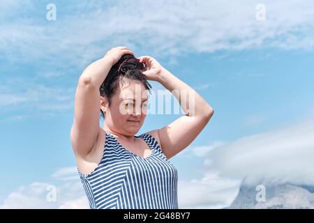 Junge asiatische Frau styling ihre schwarzen Haare mit Dreadlocks mit ihren Händen. Halblanges Porträt gegen einen bewölkten Himmel. Stockfoto