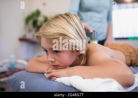 Nahaufnahme eines Jungen, der auf dem Bett liegt, während eine Therapeutin mit einem Ultraschallgerät zugeht Stockfoto