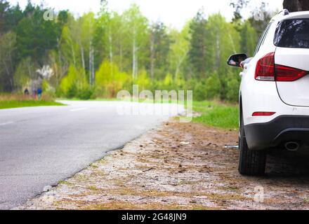 Weißer SUV auf leerer Asphaltstraße in schönem Frühlingswald geparkt. Eine Straße zwischen grünen Bäumen an einem sonnigen Frühlingstag. Reisen mit dem Auto durch einen einheimischen c Stockfoto