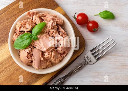 Draufsicht auf Thunfischkonserven in einer Schüssel, Gabel und rote Kirschtomaten auf einem weißen Holztisch. Gesunde Ernährung Snack von konservierten Thunfisch und frischem Gemüse. Stockfoto