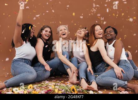 Gruppe von glücklichen Frauen auf dem braunen Hintergrund sitzen, während Blütenblätter von Blumen fallen. Lachende Weibchen verschiedener Rassen und Altersgruppen unter Blütenblättern aus Stockfoto